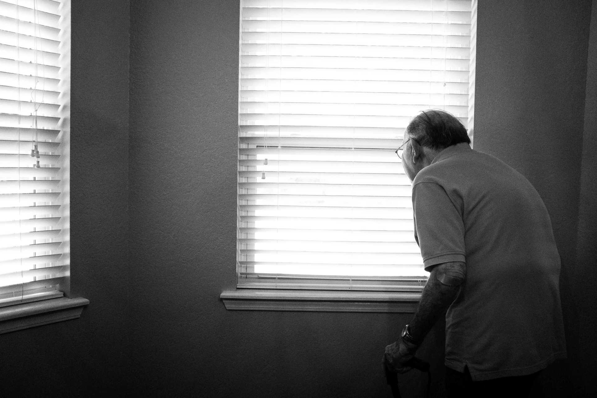Picture of man looking out of window with blinds closed, in a dark room