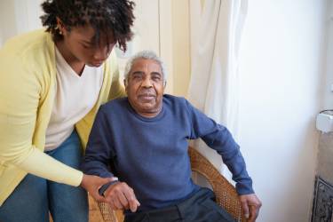 Image of man sat down in chair looking contented, with a younger woman helping him and holding his hand