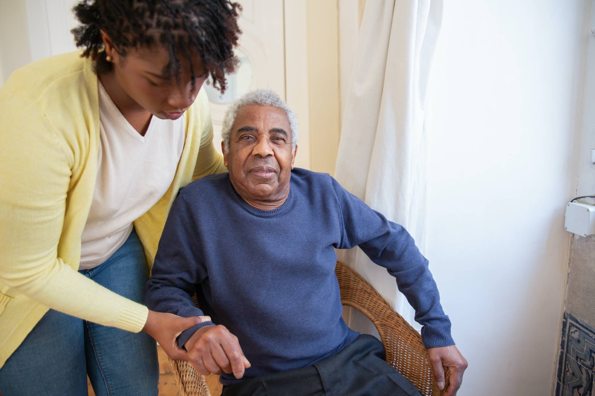 Image of man sat down in chair looking contented, with a younger woman helping him and holding his hand