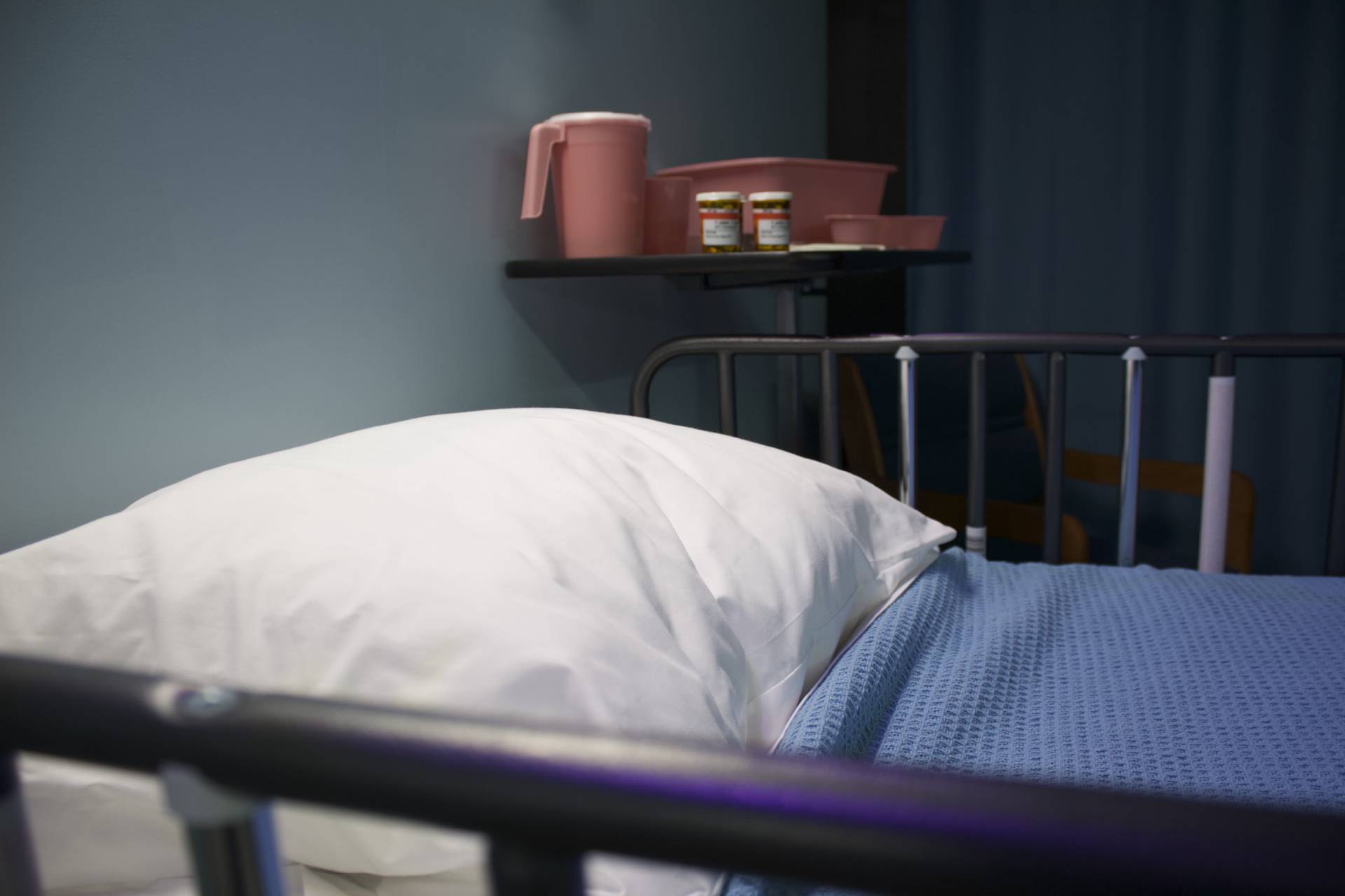 Picture of a hospital bed with a blue blanket on and white pillow, and a shelf that has a jug of water and two boxes of pills. The room is dark and there is a green curtain in the background